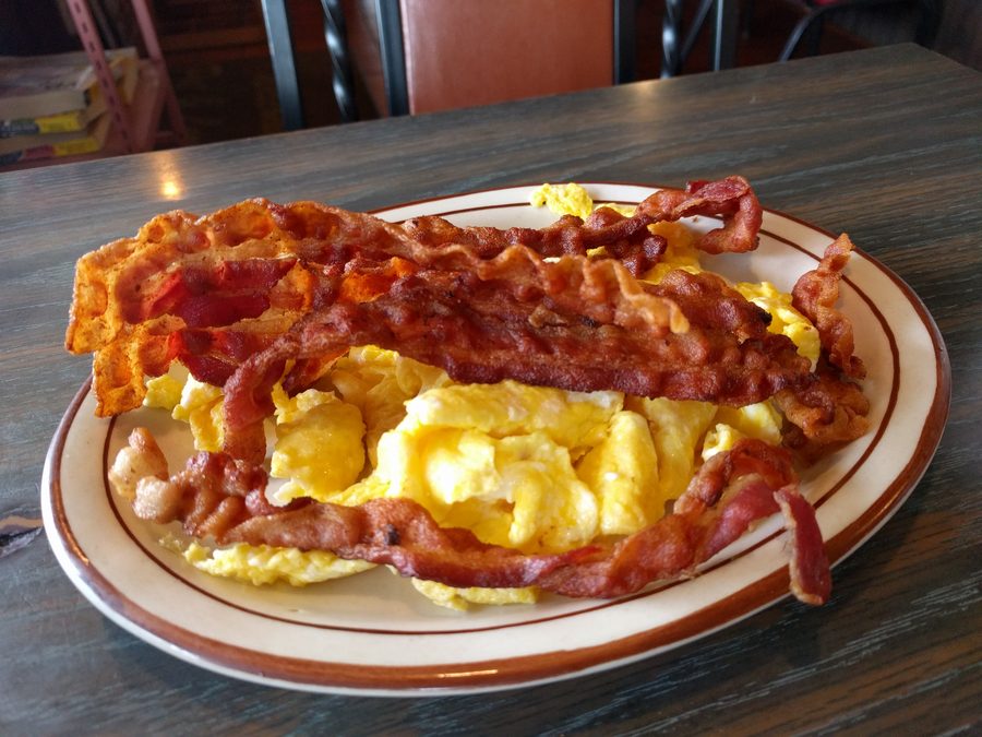 Scrambled eggs under four long strips of crisp bacon on an oval cream platter with a brown rim.