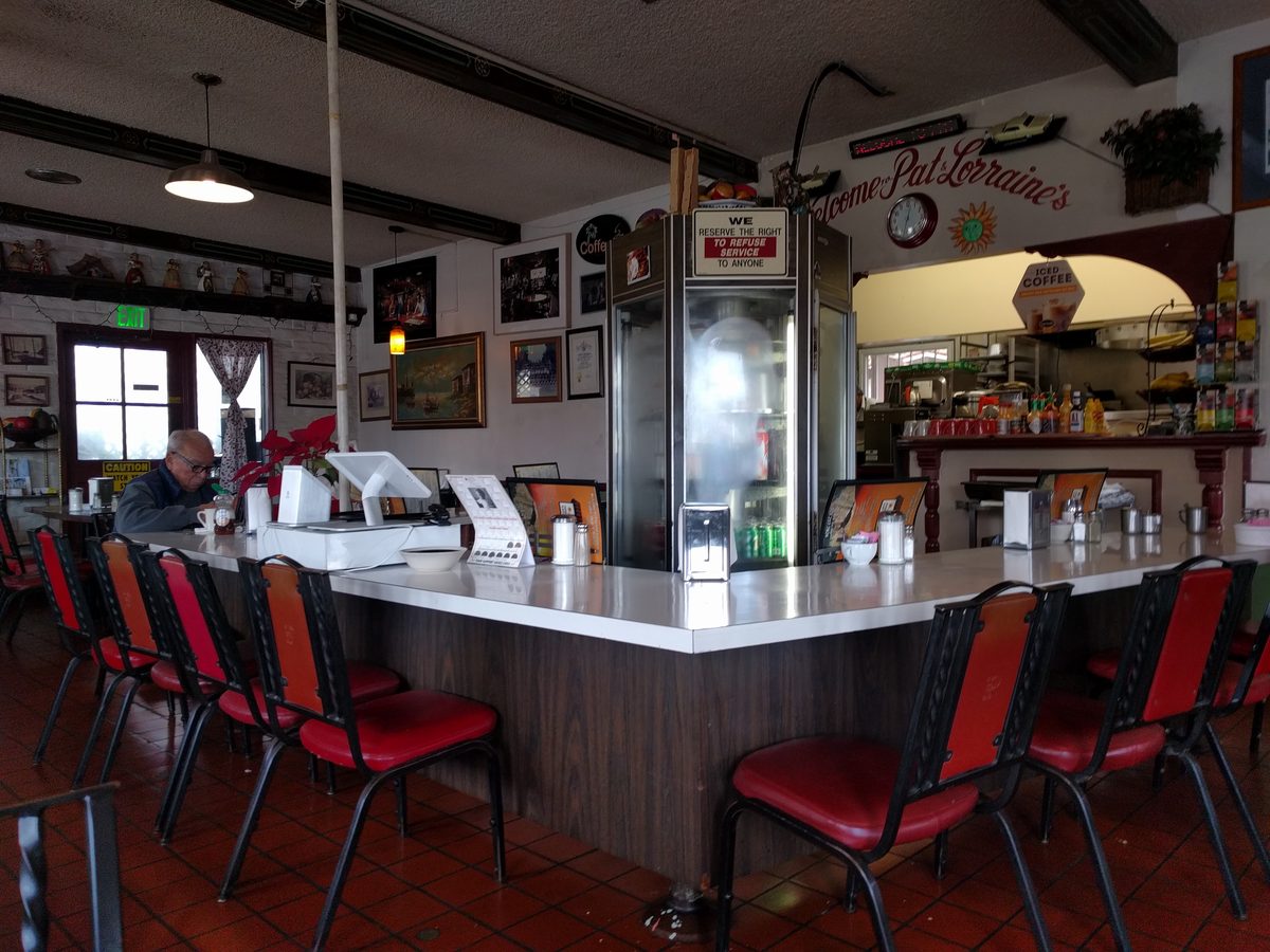 The diner counter with red vinyl stools along it, a tiled floor, and framed pictures on the walls behind.