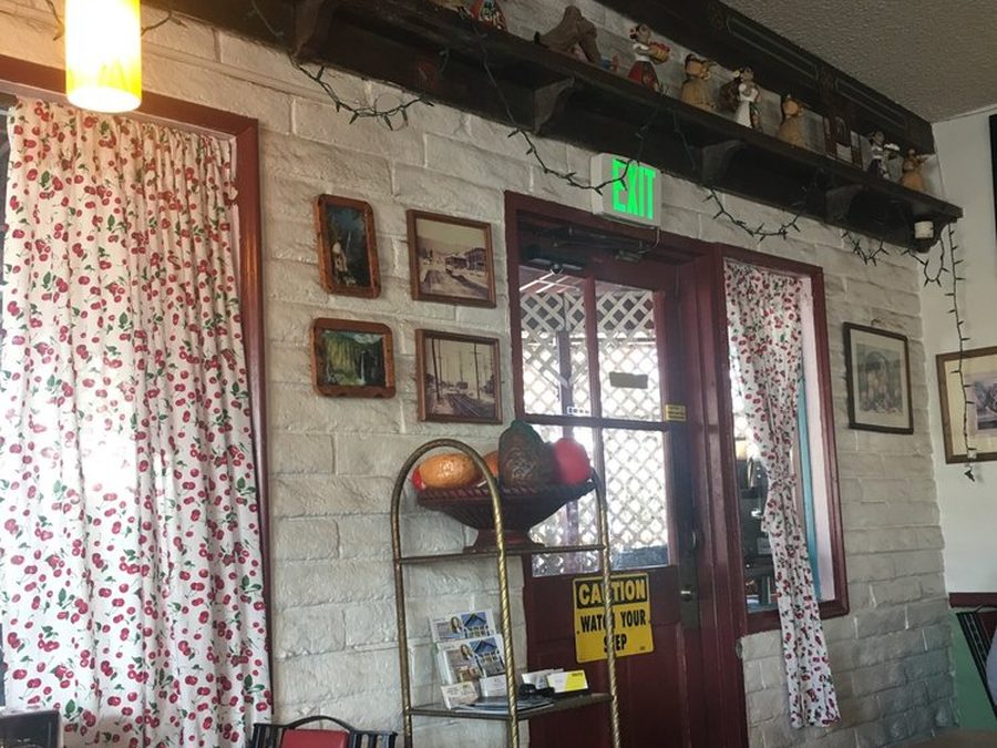 A corner of the dining room: white painted brick, cherry-print curtains at the window, small framed paintings, and a red wood door with a lattice panel.