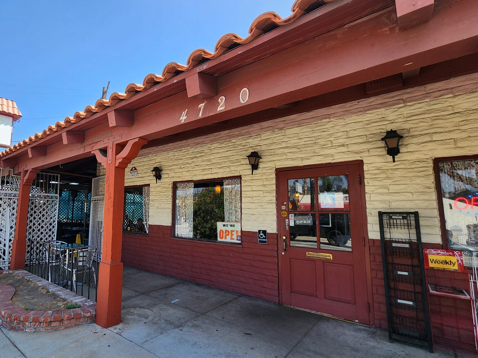 The front of the diner in daylight: a terracotta timber arcade under a red tile roof, the street number painted on the beam, and a red door beside a curtained window with a WE'RE OPEN card in it.